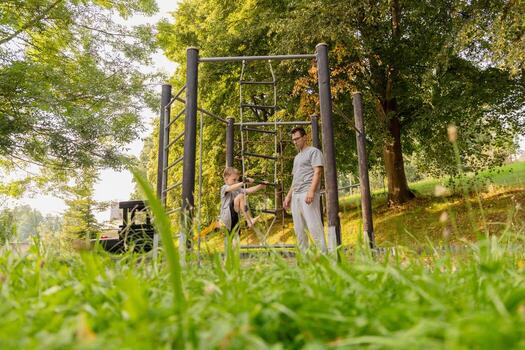 un padre y su joven hijo son teniendo divertido juntos a un patio de recreo rodeado por arboles ellos son jugando en alpinismo estructuras, creando alegre recuerdos en naturaleza foto