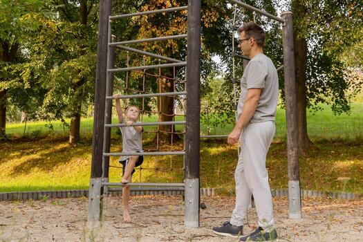 un padre relojes su hijo navegar un alpinismo estructura a un patio de recreo rodeado por arboles ellos compartir un alegre momento explorador y unión en el Fresco aire foto