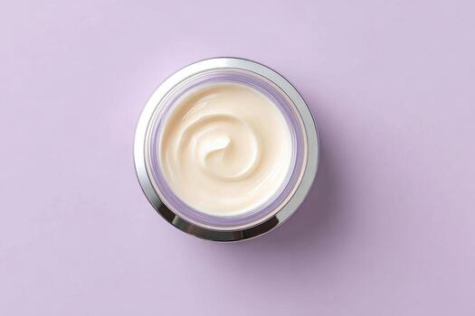 Top-down view of a round, clear jar of cream. Cream swirls in a spiral pattern. Silver metallic rim. Purple background photo
