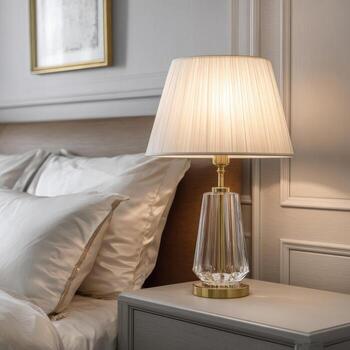 A bedside table lamp with a cream-colored pleated shade, sitting on a light-gray nightstand next to a bed with white linens photo