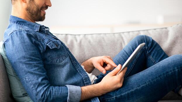 Happy middle-aged man in casual reclining on couch at home, using digital tablet, copy space, closeup. Handsome bearded man reading blog or playing games on newest pad, having fun on weekend photo