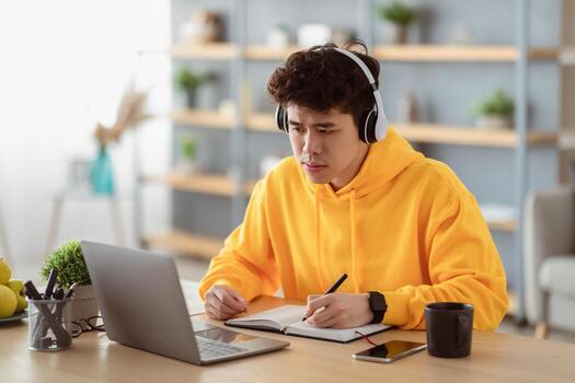Portrait of focused asian man in headset and bright yellow hoodie sitting at desk, using pc writing in notebook, taking notes, watching tutorial, lecture or webinar, studying online at home photo