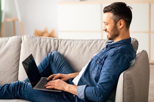 lado ver de de edad mediana hombre descansando en sofá en vivo habitación, utilizando el más nuevo computadora portátil, Copiar espacio. contento barbado hombre teniendo divertido a hogar, comprobación social medios de comunicación o chateando con amigos en computadora foto