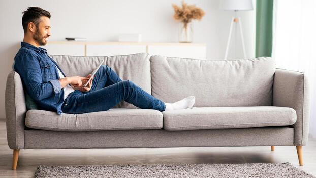 hermoso barbado hombre utilizando digital tableta mientras descansando a hogar, lado vista, Copiar espacio. relajado de edad mediana hombre reclinable en sofá en vivo habitación, acecho película o utilizando móvil aplicaciones foto