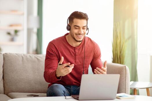 Smiling young arab man working from home, having conference with clients or colleagues, sitting on couch in front of coffee table with laptop and using headset, gesturing, copy space photo