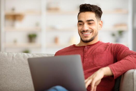 Closeup of happy middle-eastern man freelancer working from home, sitting on couch in living room and typing on notebook keyboard, chatting with clients or sending emails, copy space photo