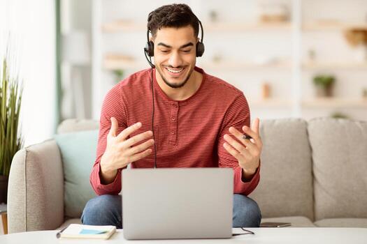 Cheerful middle-eastern guy working from home, sitting on couch in front of laptop, using headset. having conference with clients or colleagues, gesturing and talking, copy space photo