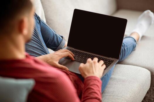 Arab guy using modern laptop with empty screen at home, mockup. Unrecognizable middle-eastern millennial man reclining on couch and using notebook with blank display, over shoulder shot photo
