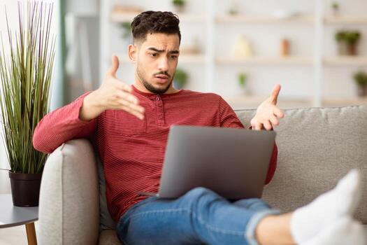 Angry arab guy using laptop at home, sitting on couch in living room, looking at notebook screen and gesturing, computer software showing error, middle-eastern man gamer lost, copy space photo