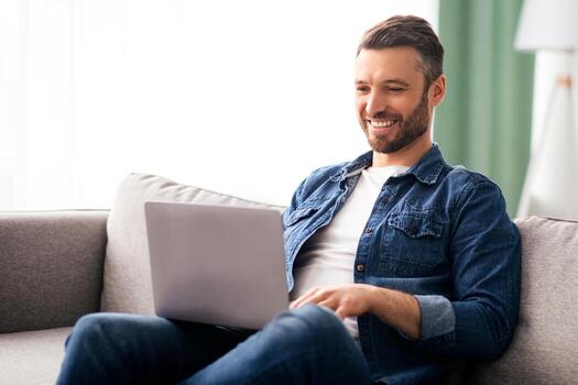 Smiling middle-aged man using modern laptop, having part-time job at home, copy space. Happy bearded man watching movie or chatting on notebook, resting on sofa in living room photo