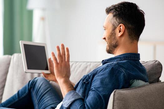 Smiling bearded man waving at digital tablet with empty screen while resting on couch at home, side view, copy space. Happy middle-aged man having conference with friends or relatives photo