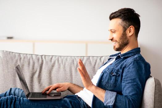Smiling middle-aged bearded man having call with friends or lover, using laptop, looking at computer screen, talking and gesturing, home interior, copy space, side view photo