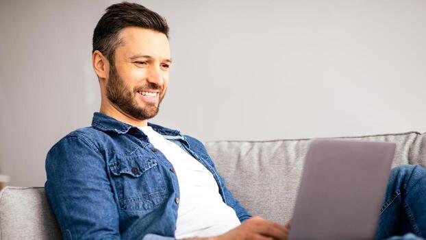 Smiling middle-aged man using modern laptop, having part-time job at home, side view, copy space. Happy bearded man watching movie on notebook, resting on sofa in living room photo