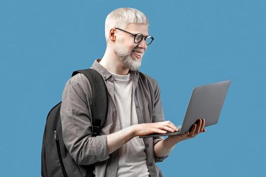 Cool guy with unusual appearance using laptop for online work or studies, wearing backpack over turquoise studio background. Happy young man communicating on web, browsing internet photo