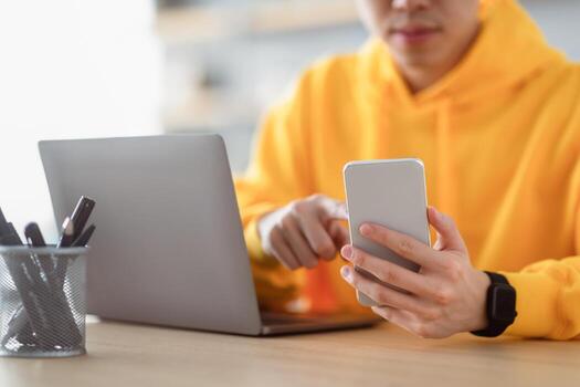 People And Technology. Closeup cropped view of unrecognizable guy in bright yellow hoodie holding smartphone using laptop computer, reading business text message, sitting at desk, selective focus photo
