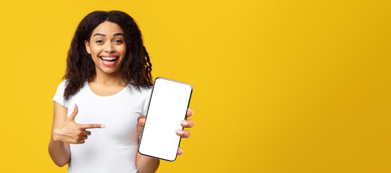 Positive black lady showing modern smartphone, pointing at empty blank screen, recommending useful mobile application, standing over yellow background, copy space, studio shot photo