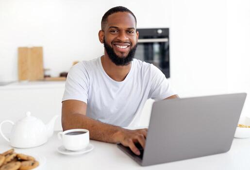 Portrait Of Smiling Black Bearded Guy Using Laptop Browsing Internet, Typing On Keyboard. Cheerful Man Having Breakfast Sitting At Dining Table In Modern Kitchen, Looking And Posing At Camera photo