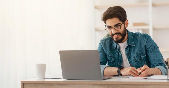 Arab businessman taking notes while working on laptop computer at home office, empty space. Freelancer noting down information from internet, planning working schedule, sitting at workplace photo