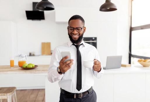 Smiling Black Business Man In White Shirt And Eyglasses Using Smartphone And Drinking Coffee While Standing At Home Office, Browsing Internet Or Social Networks, Enjoying Free Time And Break From Work photo