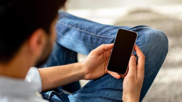 Technology concept. Over the shoulder view of unrecognizable man using cellphone with black empty screen for mockup. Guy making call or chatting online at home photo