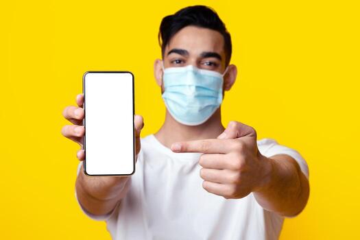 Arab guy in protective face mask showing smartphone with empty screen, middle-eastern man pointing at mobile phone with blank display, mockup, yellow background. Vaccine passport, certification photo