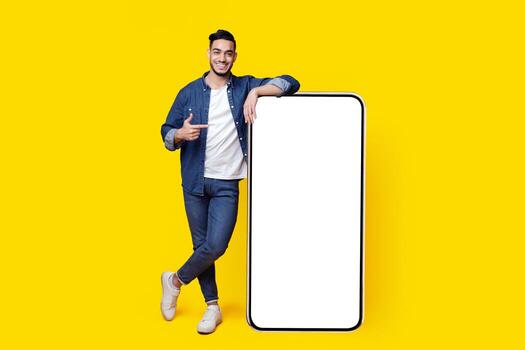 Positive arab guy with huge smartphone with blank screen posing over yellow studio background, showing thumb up and smiling, full length shot, mockup. Handsome muslim man recommending mobile app photo