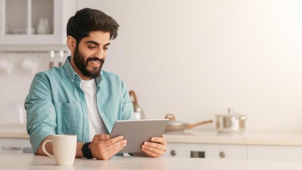 Cheerful arab man using digital tablet while drinking coffee in kitchen at home, sitting at table, enjoying morning beverage and checking social networks on tablet computer, copy space photo