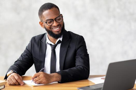 A well-dressed man with a beard sits at a desk, smiling as he takes notes during a virtual business meeting. The modern office setting features a laptop and documents scattered on the table. photo