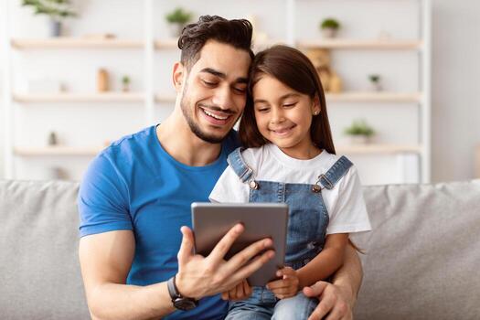 Young Father And Little Daughter Using Tablet Browsing Internet Together And Watching Movie Online Or Making Call, Sitting On Sofa At Home On Weekend. Daddy And Child Using Computer photo