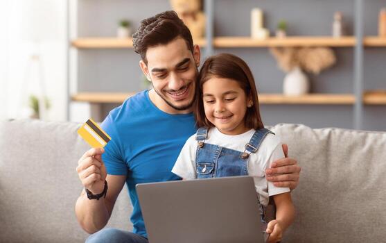 Portrait of smiling father and kid shopping together on Internet, using laptop and credit card, sitting and hugging on couch at home, looking at computer screen and smiling, order food, copy space photo