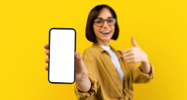I recommend this app. Excited lady holding smartphone with black blank screen, showing device and thumb up gesture, standing over yellow background. Gadget with empty space for mockup photo
