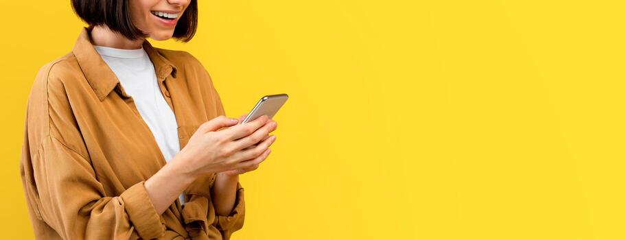 New amazing application. Happy positive young lady using smartphone and browsing internet, texting or typing sms, standing on yellow studio background, panorama with free space photo