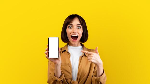 Portrait of overjoyed woman pointing at smartphone with blank screen, posing on yellow background, mockup for mobile app or website design. Surprised lady holding cellphone with empty space photo