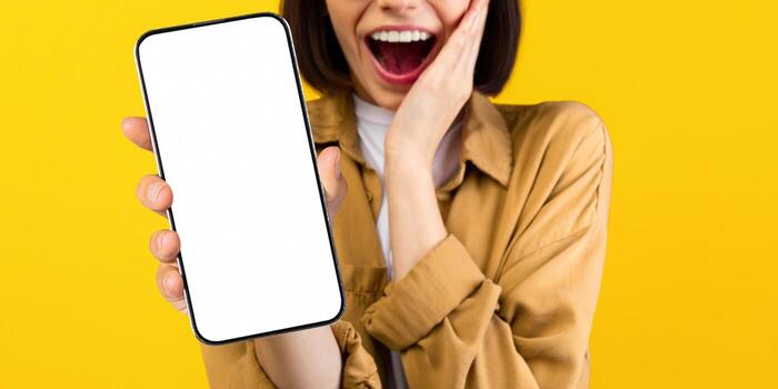 Cool app. Overjoyed woman demonstrating smartphone with blank screen, showing space for your website design or mobile application, standing on yellow background, mockup photo