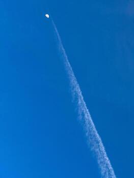 Clear blue sky with a visible moon and a straight airplane contrail rising toward it, creating a minimalistic and surreal scene. photo