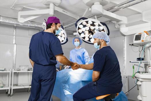Two surgeons shake hands in an operating room, with a surgical team preparing for a procedure. The scene is lit by surgical lights. photo
