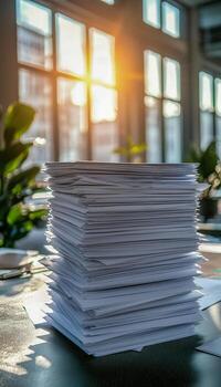 A tall stack of paper documents on a desk in a bright office with sunlight streaming through large windows photo