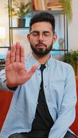 Portrait of Indian young man guy on sofa with serious expression doing stop gesture with both hands at home, warning of finish, prohibited access, declining communication, and body language danger. video