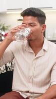 indisch Mann beim Zuhause Getränke Wasser macht groß Schluck hält Flasche und lächelt zum besser Gesundheit. arabisch Kerl auf Sofa setzt Erinnerung auf Smartphone zu trinken jeder Stunde fügt hinzu Alarm und baut Täglich Flüssigkeitszufuhr Gewohnheit video
