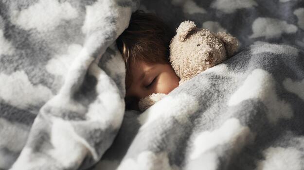 Child Sleeping With Teddy Bear Under Cloud-Patterned Blanket. Peaceful And Cozy Bedtime Scene photo