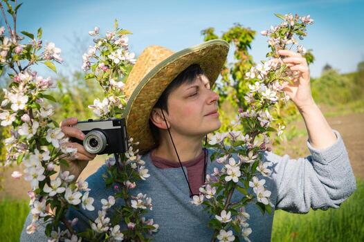 Middle aged woman in a straw hat with a vintage camera photographing a blossoming branch of an apple tree in the garden photo