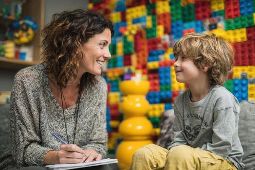 Smiling woman with curly hair engages in playful conversation with young boy in a colorful playroom filled with building blocks, fostering creativity and connection photo