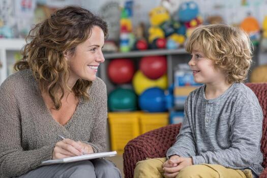 Smiling woman with curly hair engages in conversation with a young boy, in a colorful playroom filled with toys, fostering a warm and nurturing environment for learning photo