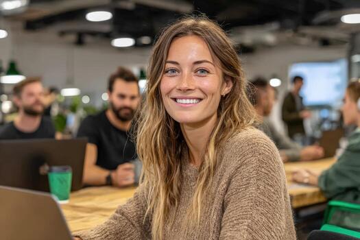 Smiling woman with long hair wearing a cozy sweater is working on a laptop in a modern office space, surrounded by colleagues engaged in collaborative tasks photo