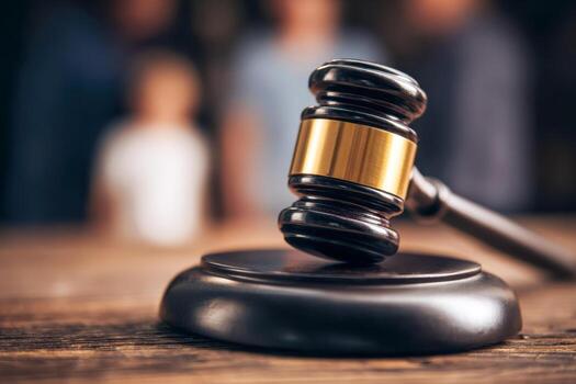 A wooden judge's gavel with a golden band resting on a matching sound block symbolizing law and justice in a courtroom setting with blurred figures background photo