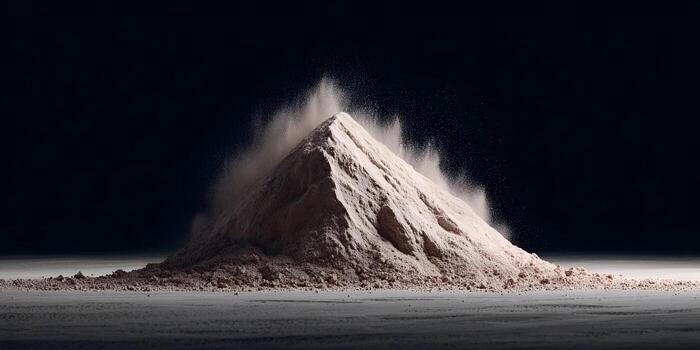 A large light brown powder pile shaped like a mountain has small particles exploding upwards from its peak against a dark background photo