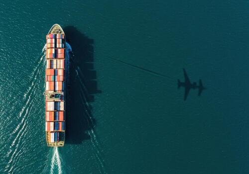 Massive cargo ship navigates deep blue ocean carrying colorful containers with airplane shadow overhead photo