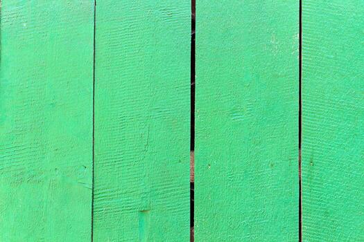 Green painted wooden fence with visible vertical planks and natural wood texture. Surface shows rough grain patterns, small cracks, and gaps between boards, creating rustic outdoor background photo