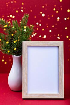 Mockup. Christmas. Empty picture frame positioned next to white vase filled with vibrant greenery on bright red background adorned with soft bokeh lights, creating festive atmosphere for holiday decor photo