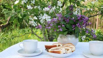 frukost i de vår trädgård. på en tabell täckt med en vit bordsduk är vit koppar med skedar, en skivad kaka och en vas av blommor. i de bakgrund är en blommande äpple träd. koppla av i de video